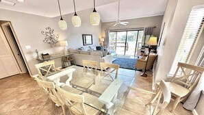 Un comedor con mesa de vidrio y sillas de madera, una sala de estar con sofá y televisor, y un ventanal con persianas.