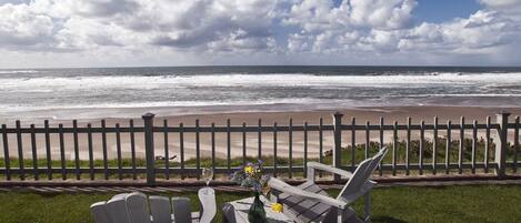 Vista de la playa con una valla de madera, dos sillas Adirondack y una mesa con un florero y una copa de vino.