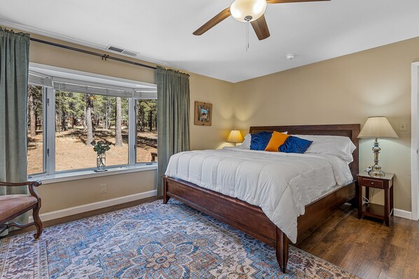 Un dormitorio con una cama grande, ventilador de techo, ventana con cortinas y vista a árboles.