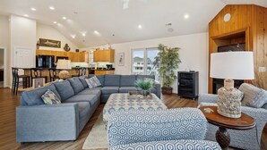 A spacious living room with a grey sectional sofa, a patterned ottoman, and a wooden coffee table. A ceiling fan and recessed lighting are visible. - SEMI Oceanfront!  Beautiful decor, ocean vws! Noon check in! Pool, HotTub (Corolla)