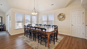A dining room with a wooden table and chairs, large windows with blinds, a ceiling light, and a door with a mirror. - SEMI Oceanfront!  Beautiful decor, ocean vws! Noon check in! Pool, HotTub (Corolla)