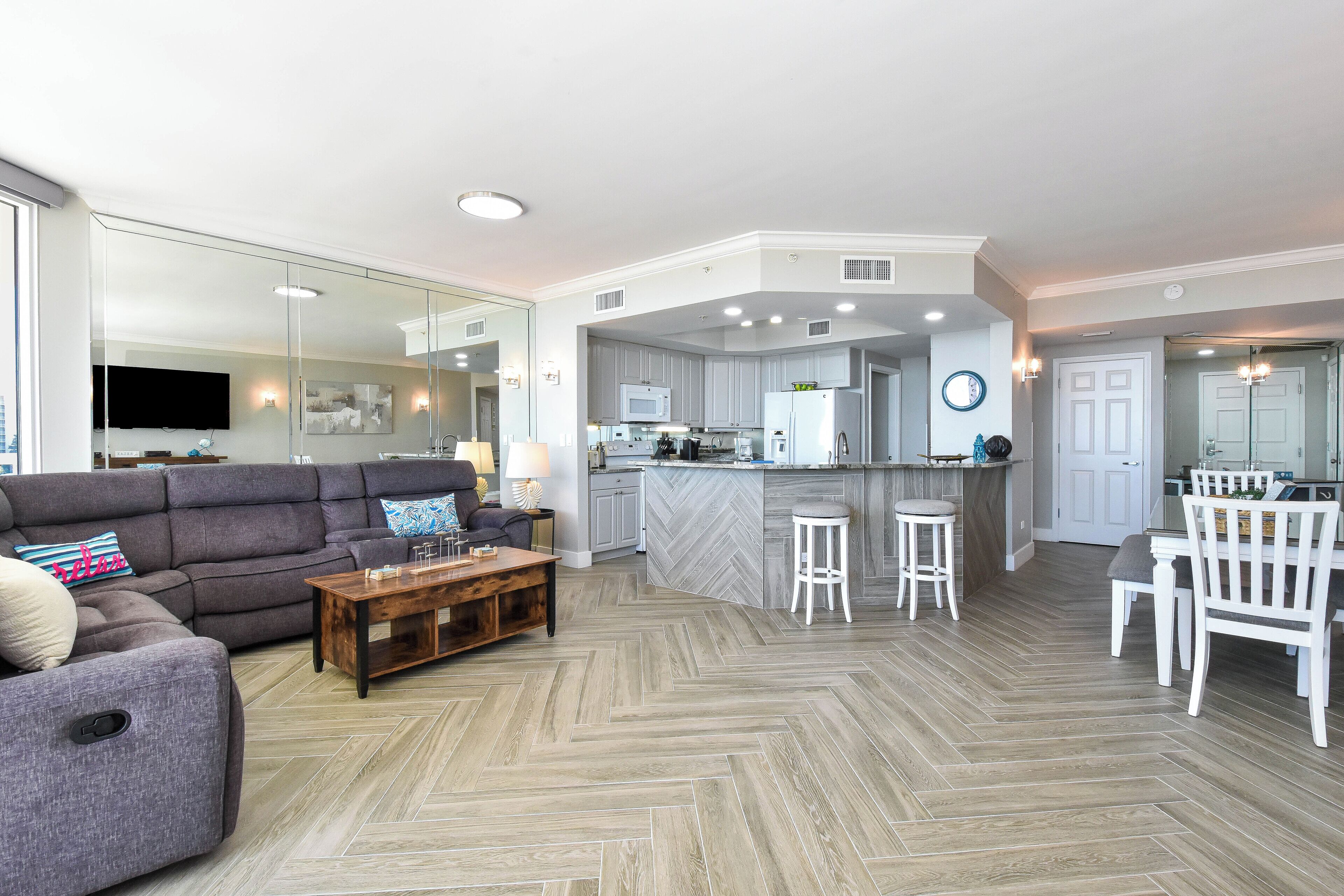 Een moderne woonkamer met een grijze modulaire bank, een houten salontafel en een visgraatvloer. Achtergrond toont een keuken met een visgraat spatwand en witte kasten.