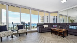 Un salón espacioso con un sofá gris, sillones estampados y una mesa de centro de madera. Grandes ventanales ofrecen vistas a la ciudad.