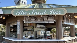 A beach-themed bar with a sign reading "The Sand Bar," a menu board, and a counter with stools. - ⛱️King in Master~Gulf View~Pools~Hot tub~Spa~2BR St. Lucia 1103 (Destin)