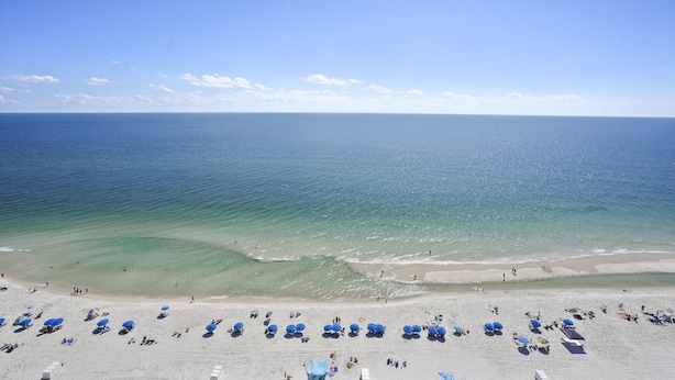 Une plage à l’eau d’un bleu limpide, avec un rivage sablonneux et de nombreux parasols bleus.