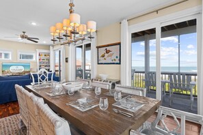 Een eetruimte met een houten tafel gedekt voor zes personen, omringd door rieten stoelen, een kroonluchter en uitzicht op de oceaan.