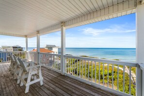 A covered wooden deck with white railings overlooking a beach with houses in the background.