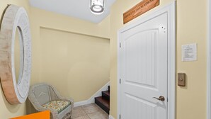 Un couloir avec une porte blanche, une chaise en osier, un grand miroir et une enseigne en bois au-dessus de la porte.