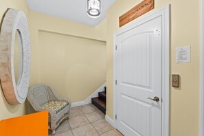 Un couloir avec une porte blanche, une chaise en osier, un grand miroir et une enseigne en bois au-dessus de la porte.