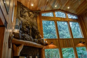 Ein rustikales Interieur mit Holzwänden, einem Steinkamin und großen Fenstern mit Blick auf dichten Wald.