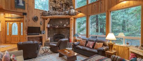 A cozy log cabin interior with a stone fireplace, leather furniture, and large windows offering forest views.