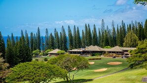 A golf course with a clubhouse, surrounded by tall pine trees and a clear blue sky. - Celebrated Villa, stylish and stunning golf views (Lahaina)