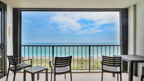 A balcony with a view of the beach, two chairs, and a table.
