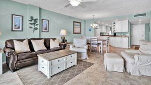 A spacious living room with a brown leather couch, white armchairs, a wooden coffee table, and a dining area with a white table and chairs. A ceiling fan and pendant lights are visible.
