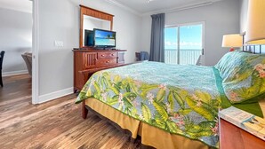 Une chambre avec un lit, un téléviseur posé sur une commode en bois, un ventilateur de plafond et une vue sur l’océan par la fenêtre.