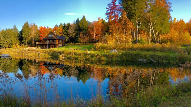 Un chalet doté d’une grande terrasse, entouré d’un étang et d’un paysage aux couleurs automnales.