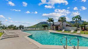 A swimming pool with lounge chairs and a small building, surrounded by palm trees and mountains.