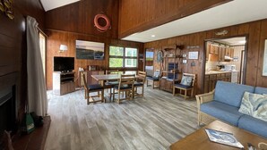 Un vaste espace de vie doté d’un plancher en bois, d’une table à manger avec des chaises, d’un canapé et d’une cuisine visible à travers une porte ouverte.