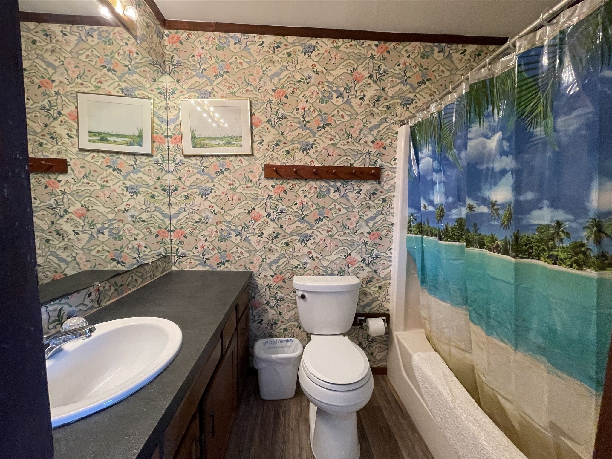 Une salle de bain avec un papier peint à motifs floraux, un lavabo, des toilettes et un rideau de douche représentant une scène tropicale.