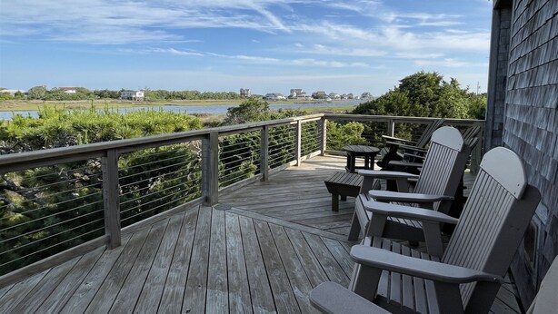 Une terrasse en bois, agrémentée de chaises Adirondack, qui offre une vue sur une étendue d’eau et sur des bâtiments situés au loin.