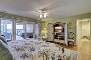 Une chambre avec un lit, une télévision et un ventilateur de plafond. Une porte-fenêtre coulissante donne accès à un balcon aménagé avec du mobilier d’extérieur.