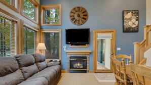 A living room with a fireplace, a large clock, and a television on the wall. - 3BR Slopeside Disciples Village Condo 604 - Ski In/Ski Out on Boyneland! (Boyne Falls)