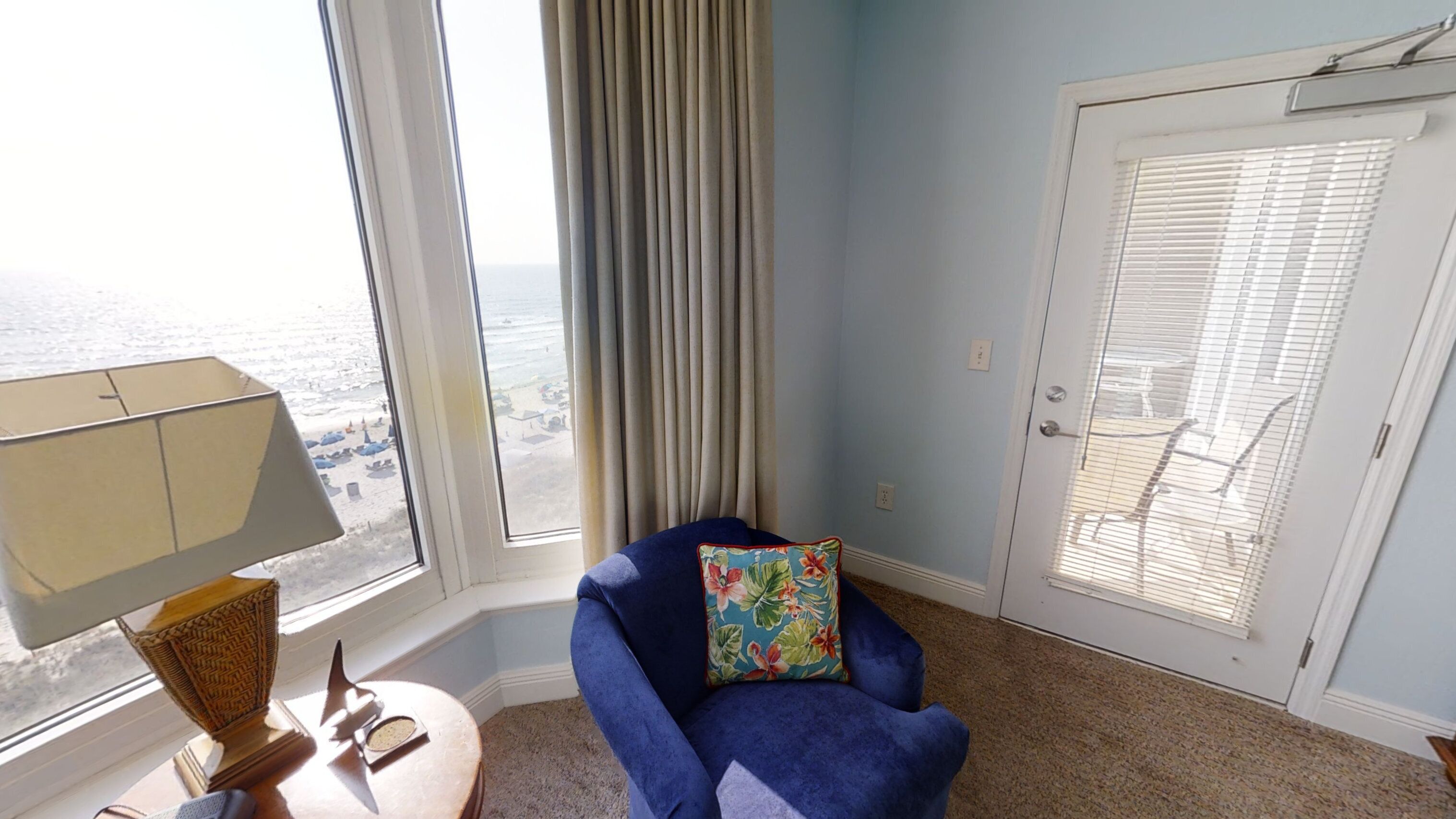 Ein Zimmer mit Meerblick, ein blauer Sessel mit floralem Kissen, eine Lampe und eine Tür mit Jalousien.