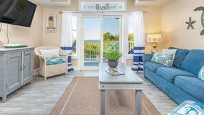 A living room with a blue sofa, a wicker chair, a television, and a view of the outdoors through a glass door. - Bay View Villas 107 | Waterfront, Community Pool (Tybee Island)