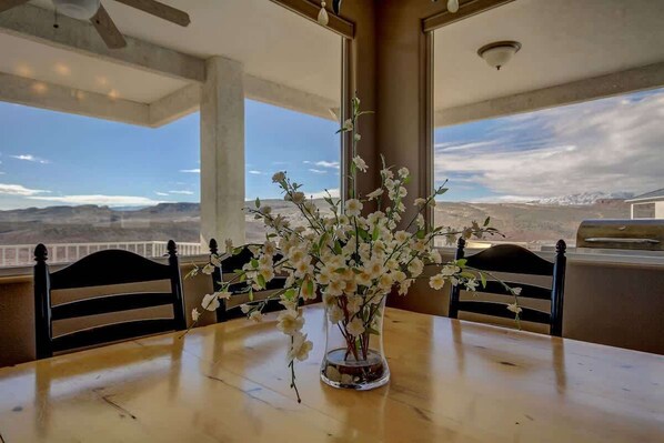 A dining table with a vase of flowers, two chairs, and a view of a mountainous landscape through large windows.