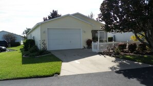 A single-story house with a garage, a front yard with grass and a small tree, and a white picket fence. - AMAZING LOCATION! Beautiful 2/2 Patio Villa w/GOLF CART & WiFi Close to Town (The Villages)