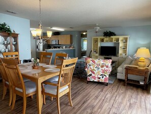 Un comedor con sillas y mesa de madera, una sala de estar con sofá y televisión, y una cocina al fondo.