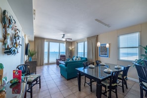 A spacious living area with a large sofa, dining table, and chairs. There is a ceiling fan, large windows with curtains, and a view of the outdoors.
