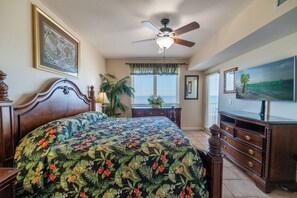 Ein Schlafzimmer mit Bett, Deckenventilator, Kommode, Fernseher und einem Fenster mit Jalousien.