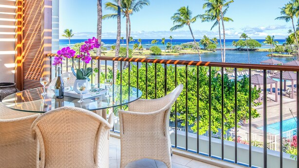 Un balcón con sillas de mimbre y una mesa de vidrio, con vistas a una piscina y un paisaje tropical con palmeras y vista al océano.