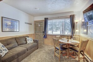 A cozy living room with a brown sofa, a wooden dining table with chairs, a framed picture on the wall, and a window with curtains.