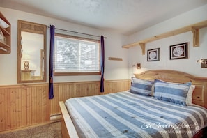 A bedroom with a wooden headboard, a large window with curtains, a bed with striped bedding, and two framed pictures on the wall.