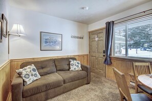 A cozy room with a brown sofa, a wooden chair, a small table, a framed picture on the wall, and a window with blinds.