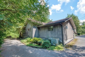 A rustic wooden cabin with a porch, surrounded by lush greenery and a clear sky.