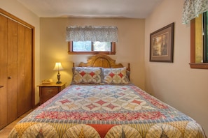A bedroom with a wooden bed, a nightstand with a lamp, a window with a floral valance, and a framed picture on the wall.