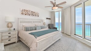Un'ampia camera da letto con un letto grande, un ventilatore a soffitto, un comodino, una sedia e vista sull'oceano attraverso ampie finestre.