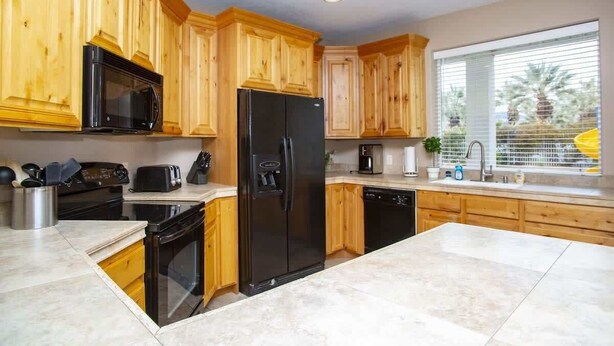 A kitchen with wooden cabinets, a black refrigerator, a microwave, a stove, and a window with blinds.