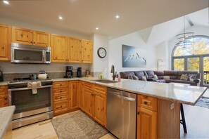 A modern kitchen with wooden cabinets, stainless steel appliances, a central island, and a view of autumn trees through large windows.