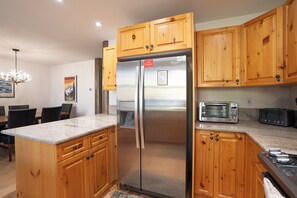A kitchen with wooden cabinets, a stainless steel refrigerator, a built-in toaster, and a dining area with a chandelier. - Elk Ridge Trail Townhome: Hot Tub, Spacious, Cool Location (Breckenridge)