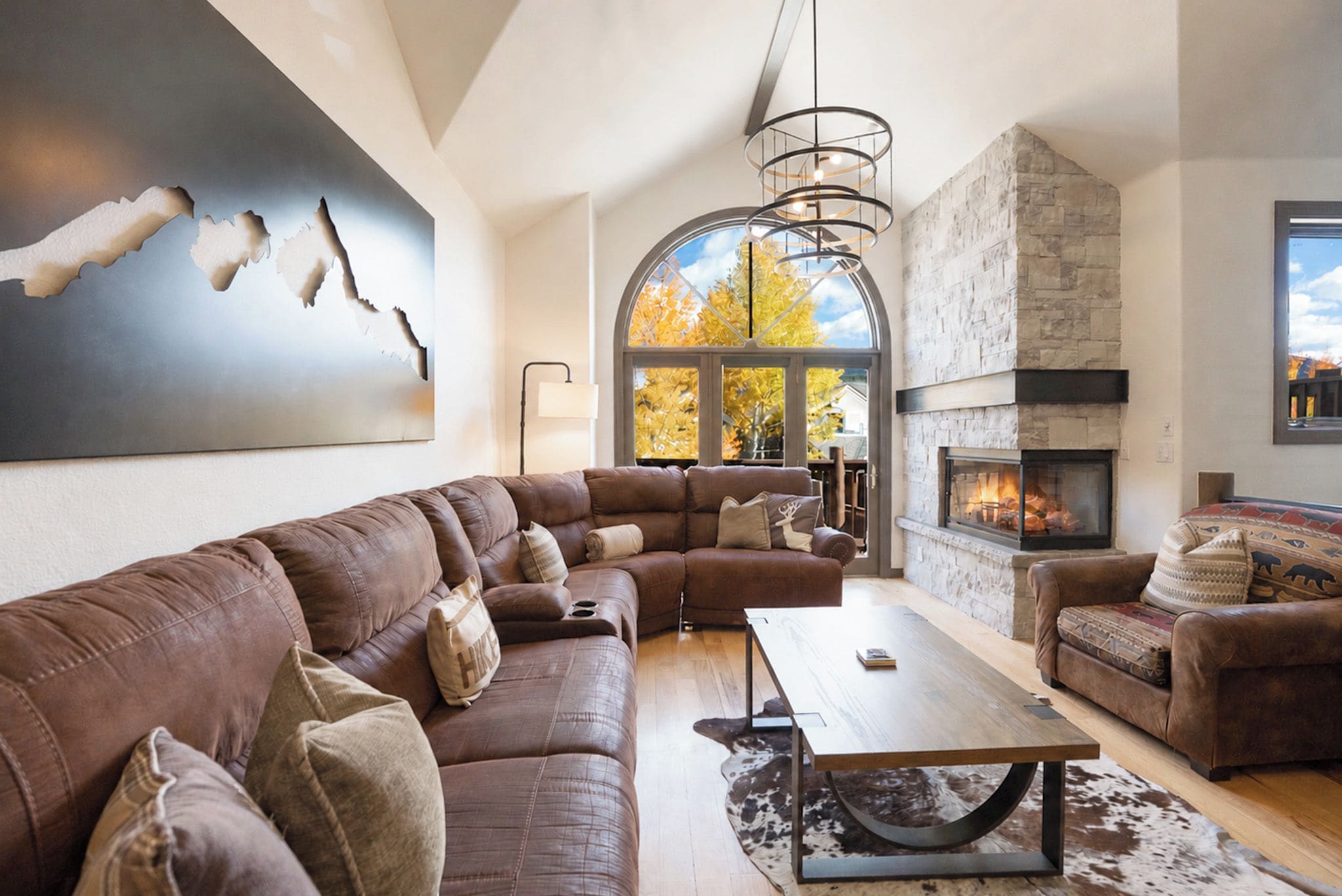 A modern living room with a sectional sofa, a fireplace, and a large window with autumn foliage.