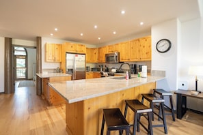 A modern kitchen with wooden cabinets, stainless steel appliances, a large island with bar stools, and a clock on the wall. - Elk Ridge Trail Townhome: Hot Tub, Spacious, Cool Location (Breckenridge)