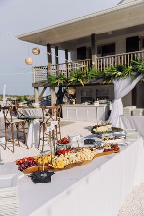 A buffet table with various food items, a white tablecloth, and a backdrop of a building with a balcony.