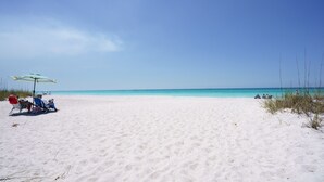 En strand med hvidt sand og klart, blåt vand. To personer befinder sig under en stribet parasol, og der er enkelte andre i det fjerne.