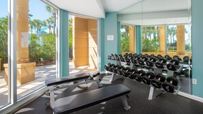 Ein Fitnessstudio mit Bänken, Kurzhanteln und einem Fenster mit Blick auf Palmen und ein Gebäude.