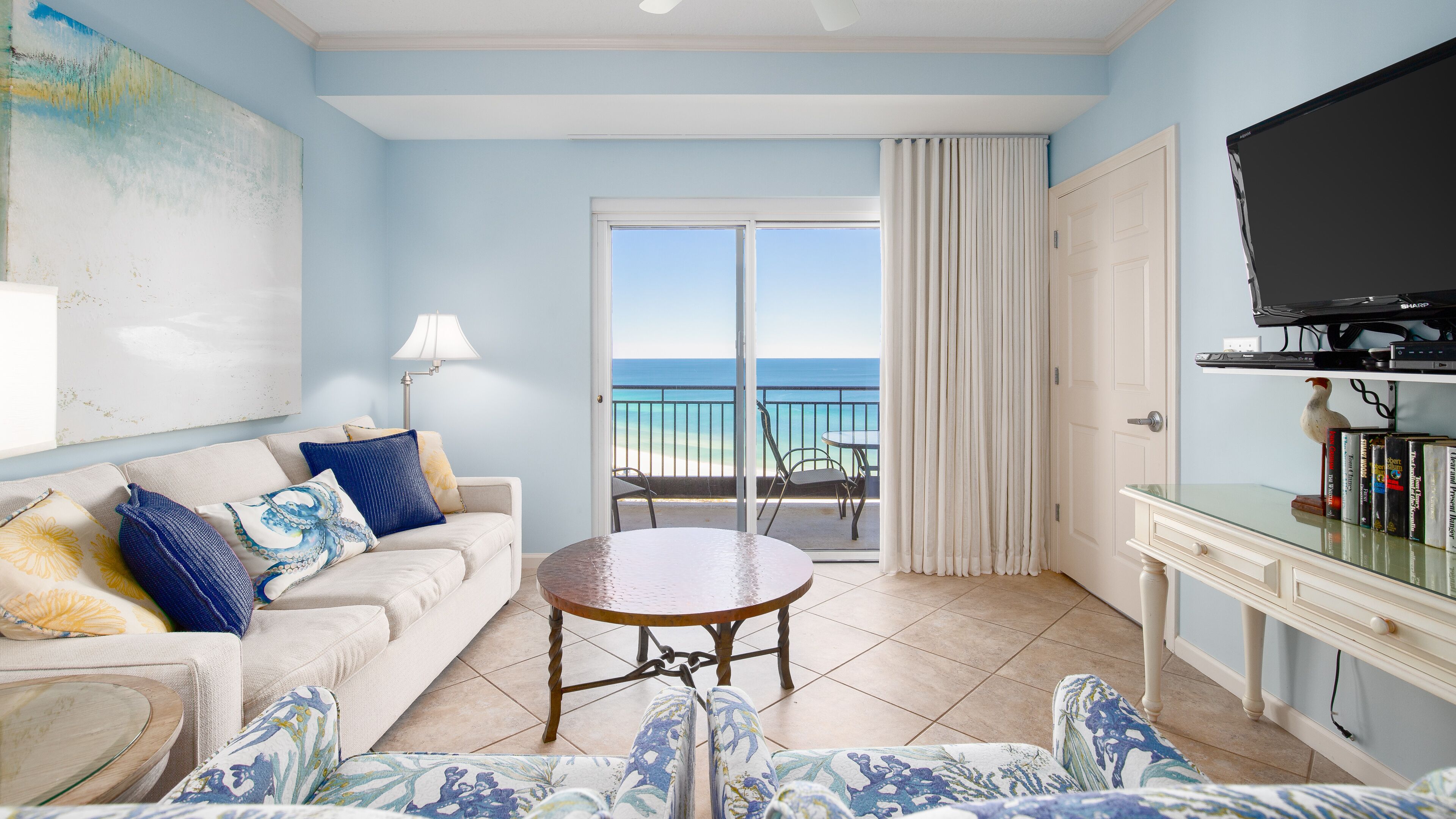 A room with a sofa, a round table, a TV, and a view of the beach.