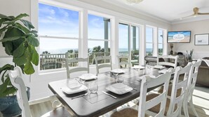 A dining area with a view of the ocean, white chairs, a wooden table, and a chandelier.